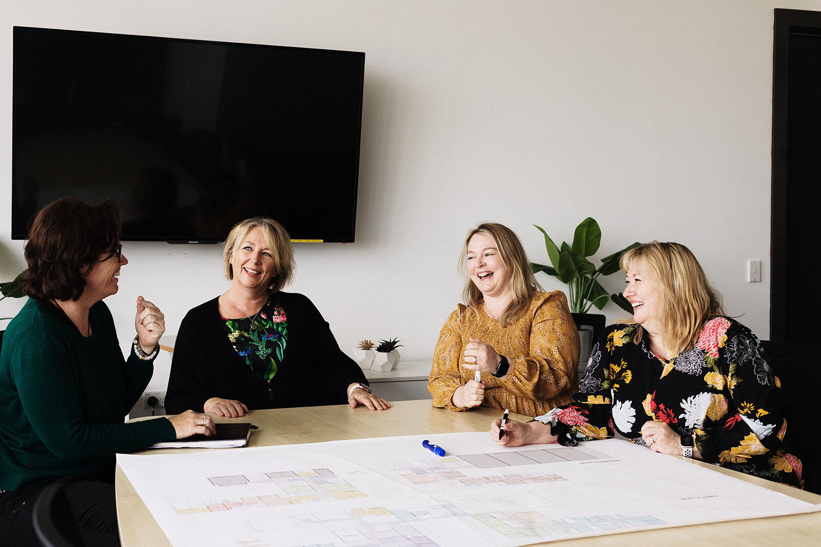 Four women seated at a meeting table smiling and talking in a modern office space.