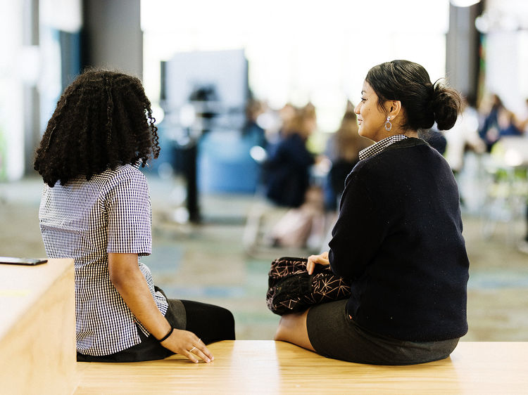 Two students sitting and chatting on a wooden bench in a bright, open school space.
