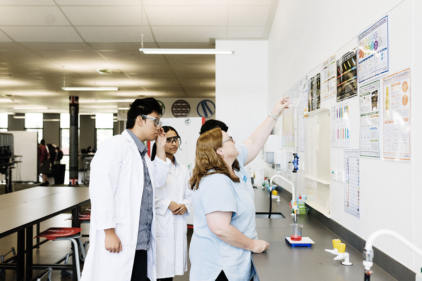 Students in science lab coats listen as a teacher explains a wall display of scientific posters and diagrams.