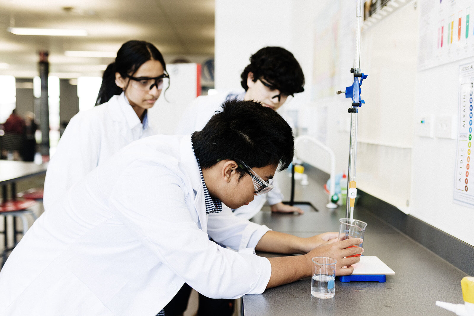 Three students in lab coats conducting a science experiment using beakers and a burette.