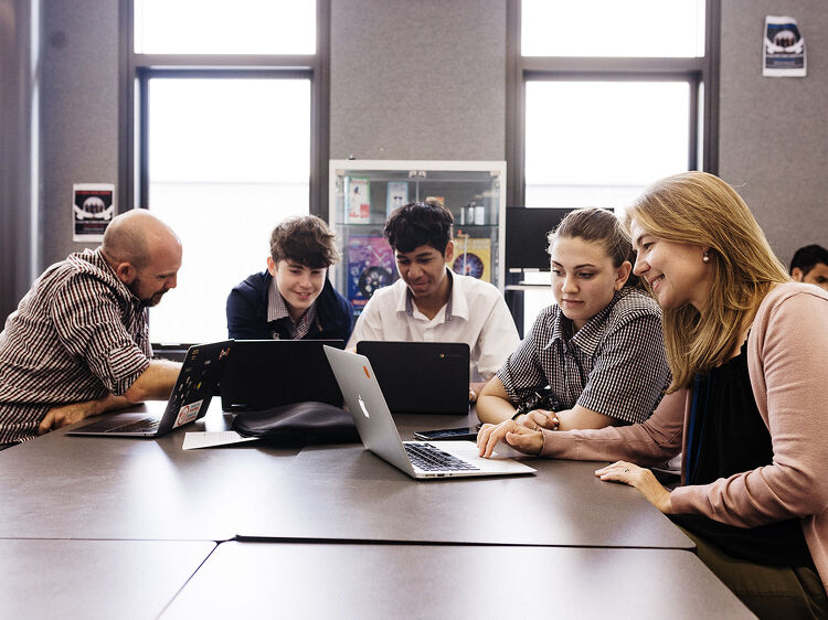 A classroom scene with two teachers and three students closely gathered around laptops, engaged in learning.