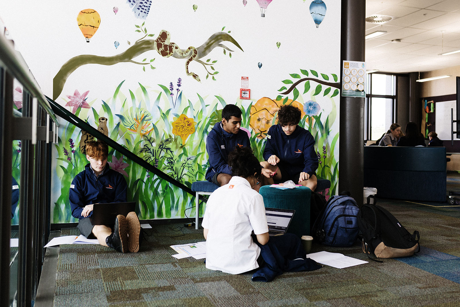 A group of students in school uniform sit on the floor under a colourful mural, working on laptops and papers in a casual learning environment.
