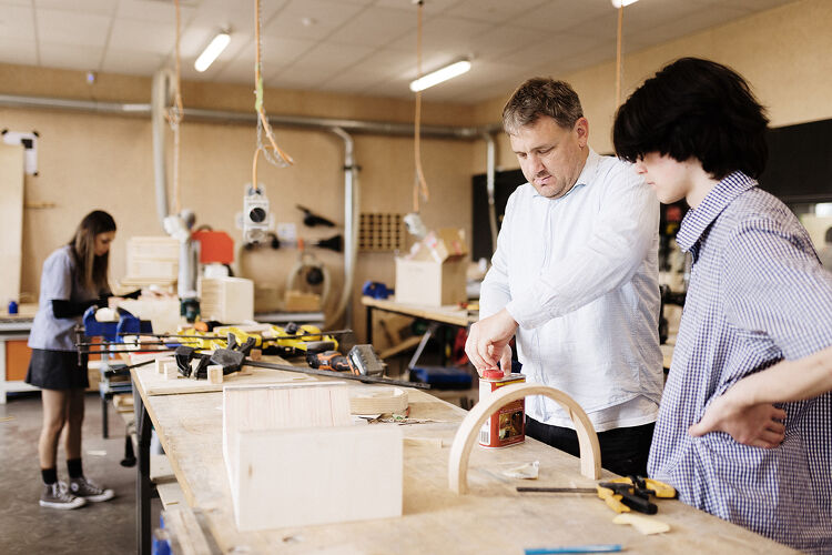 A student receives hands-on guidance from a teacher in a woodwork class while using tools on a workbench.