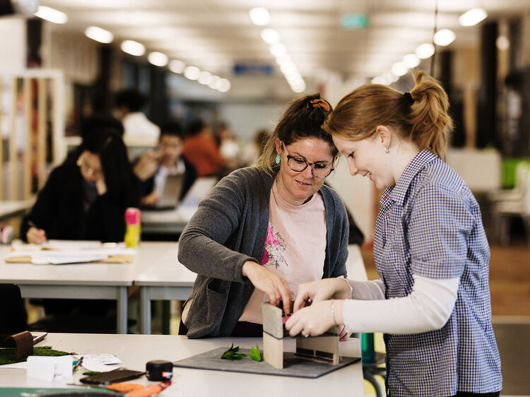 Teacher and student working together on a model in a design classroom, with others in the background.
