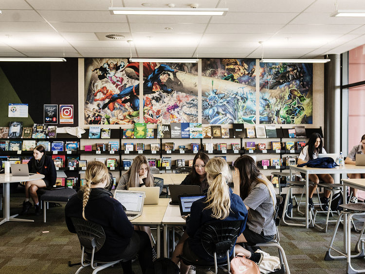 Groups of students work on laptops in the school library, surrounded by books and a vibrant superhero mural.