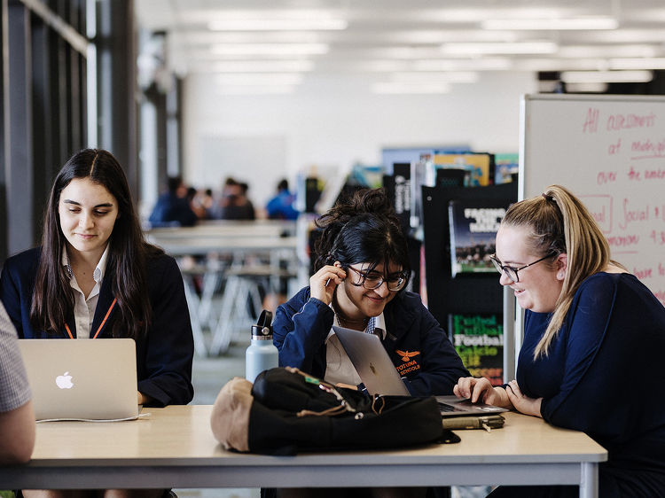 Three students sit together at a table using laptops, smiling and working collaboratively in a shared learning space.