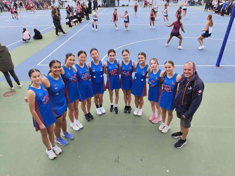 Netball team in blue patterned uniforms posing on an outdoor court with two girls kneeling in front holding a ball.