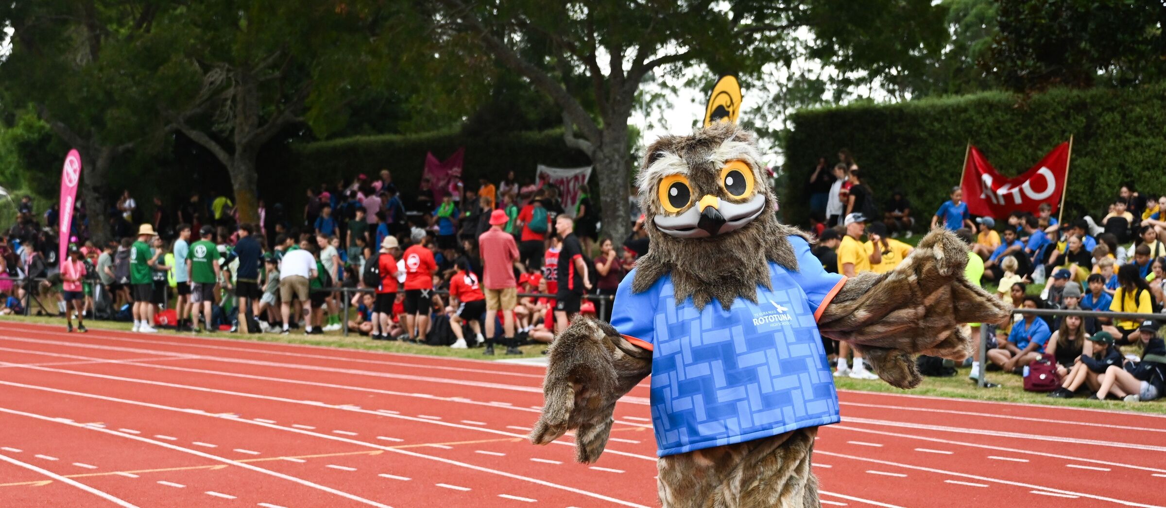 A school athletics event with a mascot standing in front of a cheering crowd near the running track.