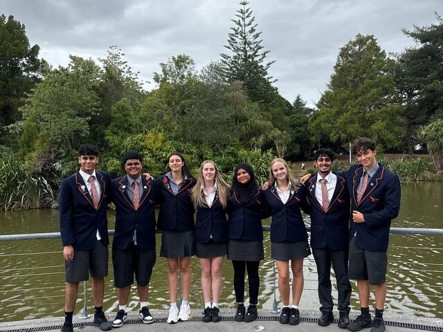 Eight senior students standing in a row smiling beside a lake in their school uniforms