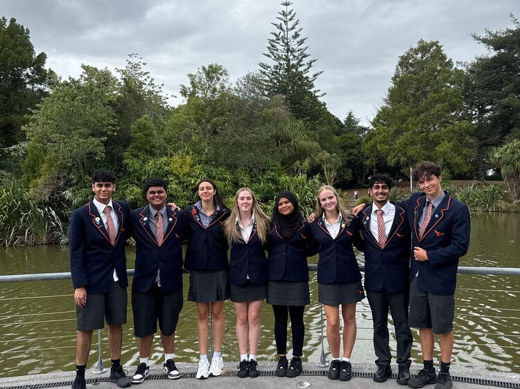 Eight senior students standing in a row smiling beside a lake in their school uniforms