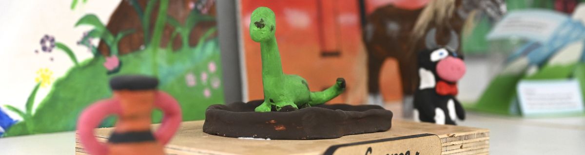 Student clay models on display, including a green dinosaur sculpture on a wooden base.