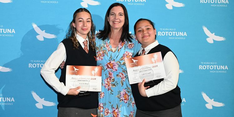 Two students holding certificates with a staff member in front of a Rototuna High School branded backdrop.
