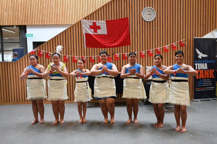 Group of students in Tongan traditional dress performing a cultural presentation in front of a Tongan flag.
