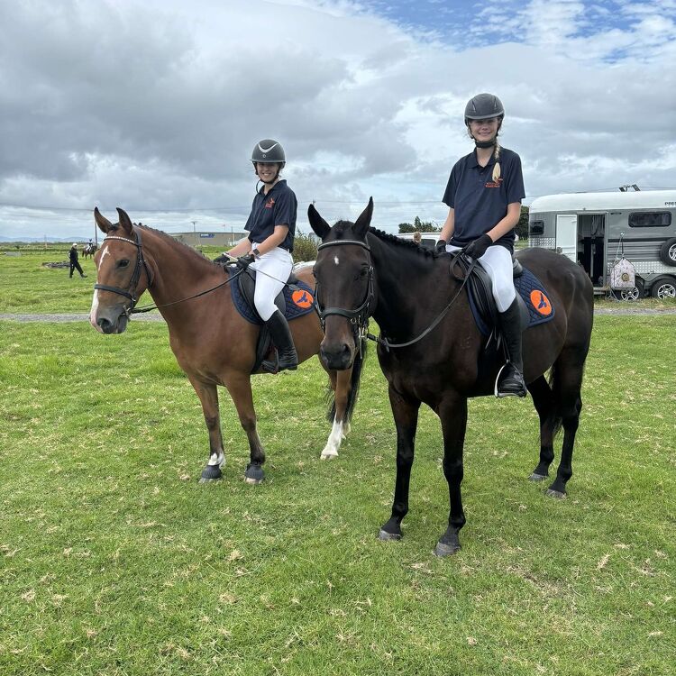 Two students in black polos and white jodhpurs sitting on horses, posing together at an equestrian event on a grassy field.