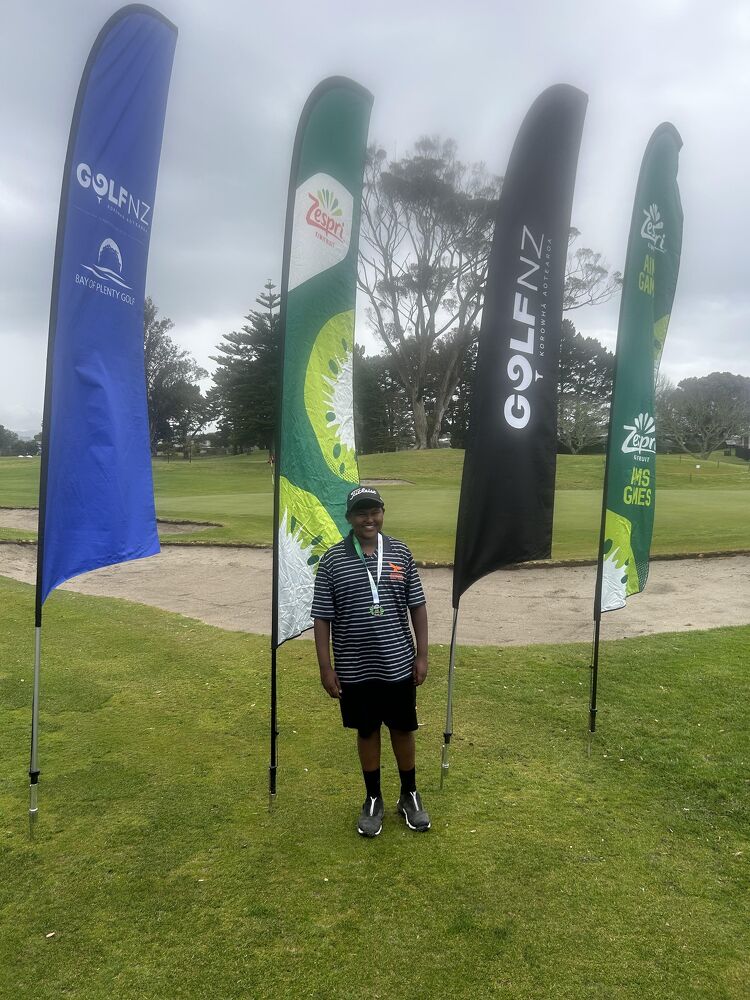 A student golfer smiling in front of golf event flags at a competition on a golf course.