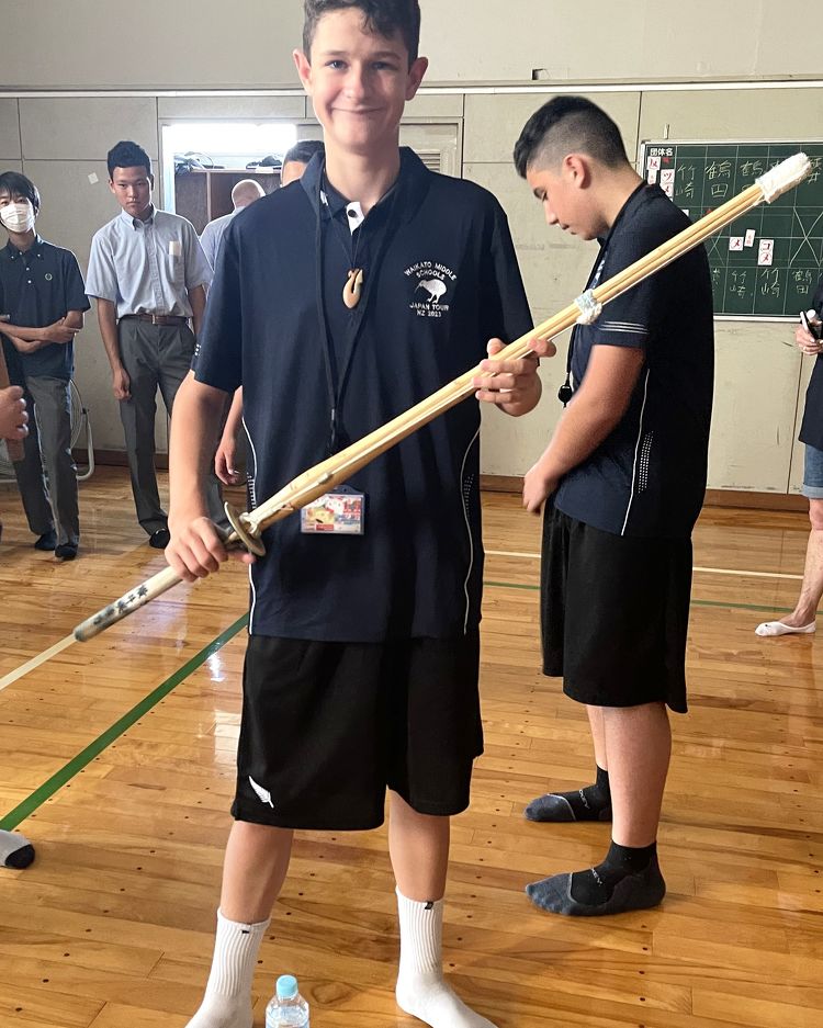 A student holds a wooden sword with confidence during a school cultural exchange or visit, surrounded by peers in a gym.