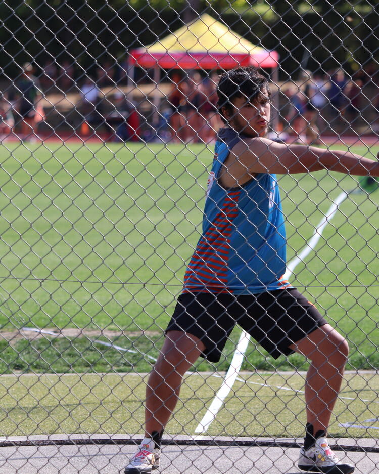 Male student in a blue and orange sports uniform prepares to throw a discus behind a chain-link fence.