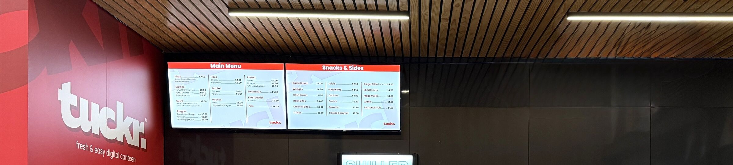 Digital menu boards and signage for "tuckr." displayed above a school tuck shop counter.