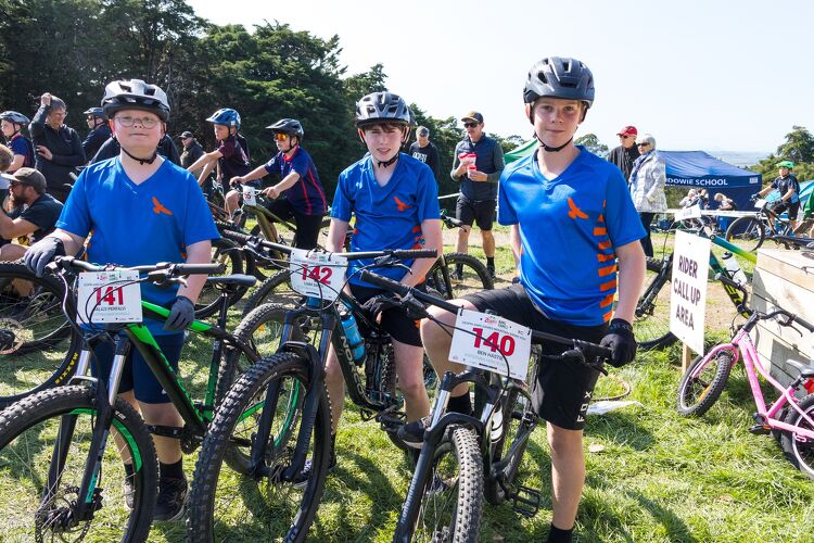 Three boys in blue sports uniforms standing with mountain bikes, wearing helmets and numbered race plates, ready for a cycling competition.