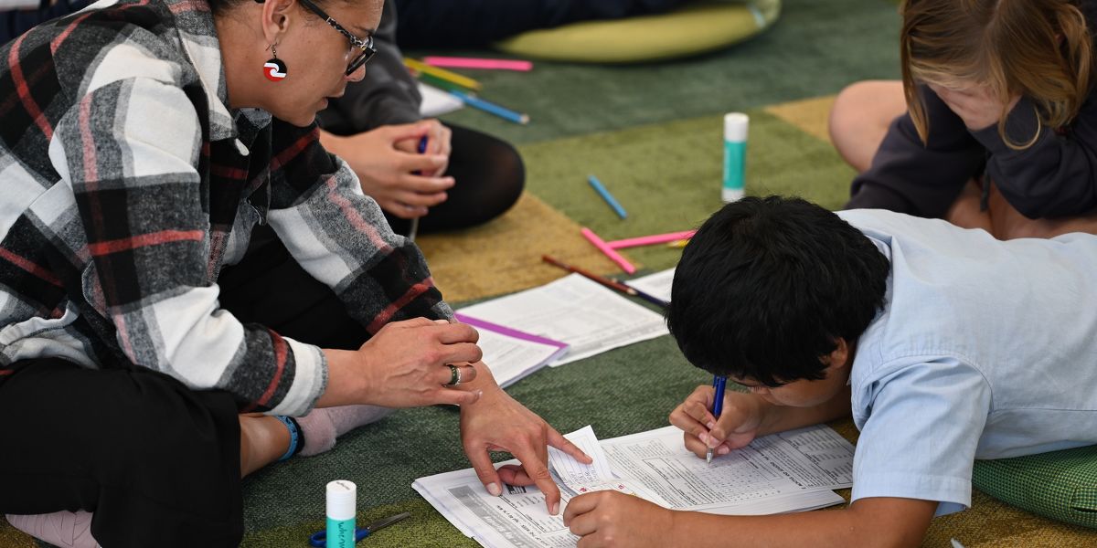 Teacher helping a student on the floor with classwork using worksheets and coloured pens during a relaxed classroom session.