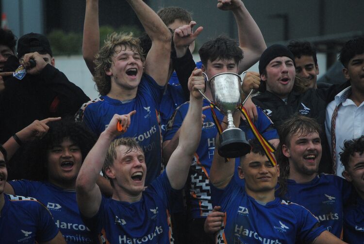 Boys rugby team celebrating and lifting a championship trophy, wearing blue Harcourts Hamilton jerseys.
