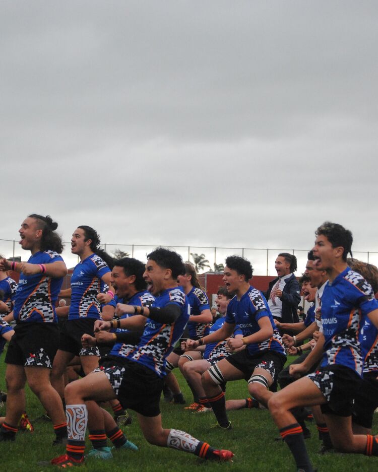 Boys rugby team performing a haka in formation on a grassy field, wearing blue and black jerseys with cultural patterns.