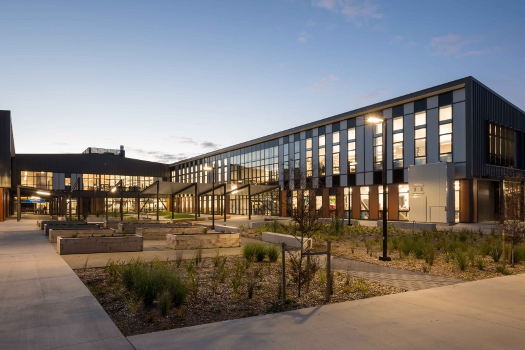 Modern school building at dusk with lights on and outdoor garden beds in front.