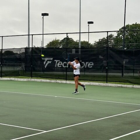 A female tennis player mid-air hitting a shot during a match on a green outdoor court.