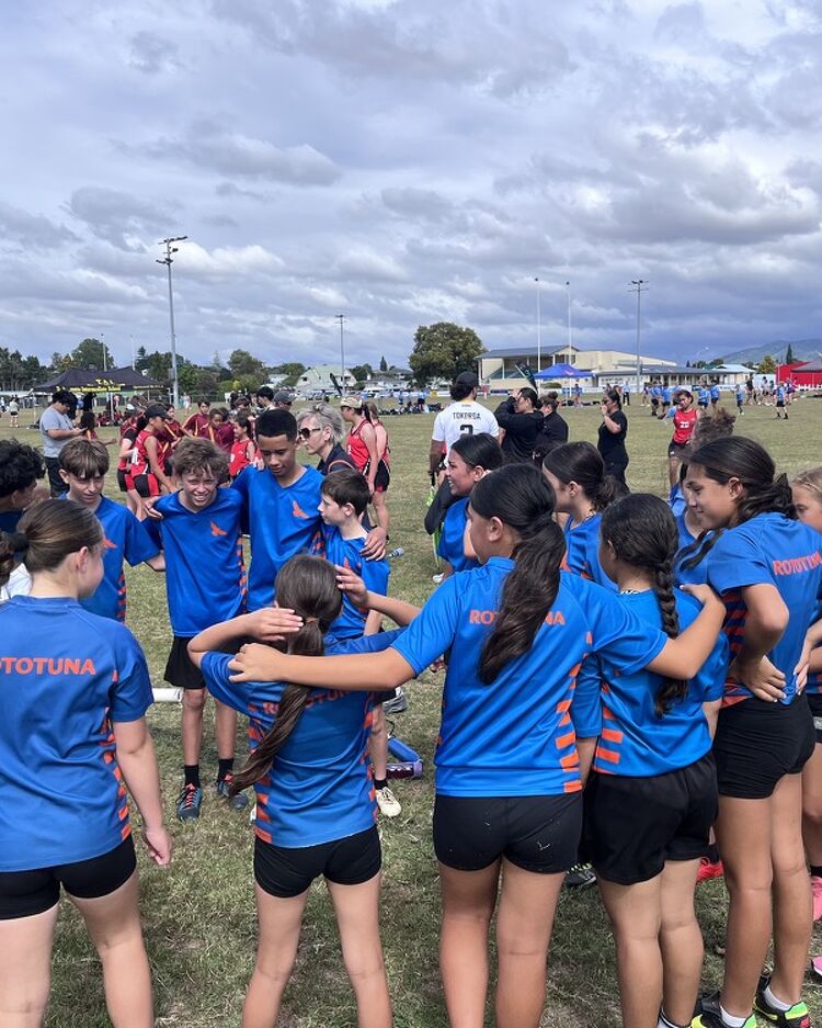 A group of students in blue Rototuna t-shirts standing in a circle with arms around each other on a sports field.