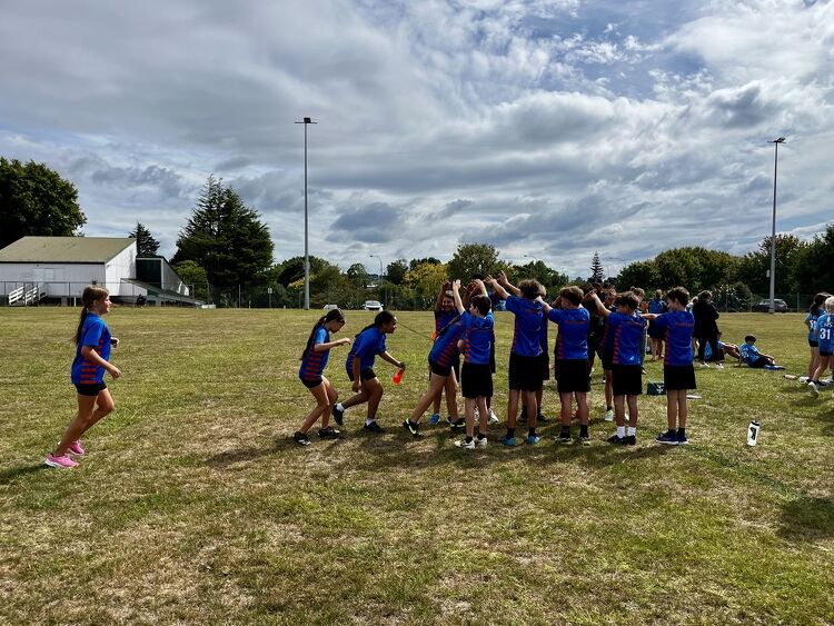 Students forming a tunnel with their arms on a school field while others run through it as part of a team activity.