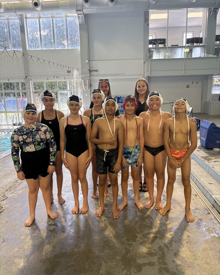 Group of young students in swimwear posing poolside with their coaches at an indoor aquatic centre.